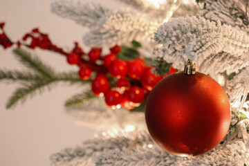 Close-up of a red Christmas ornament hanging on a snow-covered tree branch with festive berries and warm holiday lights.