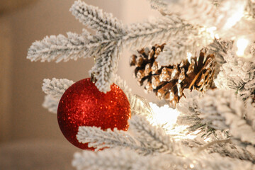 Festive close-up of a red glitter Christmas ornament and snow-covered pine cone on a decorated tree with warm holiday lights.