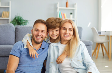 Portrait of happy united family parents and child sitting on floor and looking at camera. Joyful son hugging his smiling mother and father from behind, showing warmth and closeness. Family, love, care