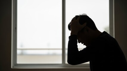 Silhouette of frustrated young asian male by window in thoughtful pose