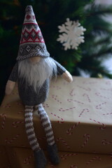 Cozy Christmas scene with a small Nordic gnome sitting on wrapped gifts under a decorated tree. Kraft paper boxes, soft handmade toy and natural holiday decor create a warm festive atmosphere at home.