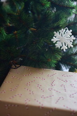 White snowflake ornament hanging on green pine branches above a kraft paper gift box with candy cane pattern. Minimal holiday style with natural textures and cozy festive mood.