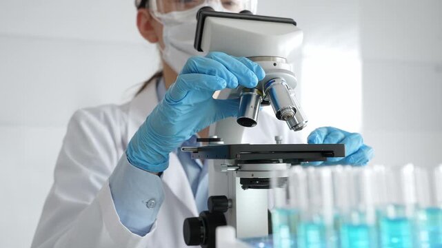Female scientist wearing protective equipment checking microscope, analyzing blue test tubes in modern laboratory research setting. Medicine, healthcare and science concept