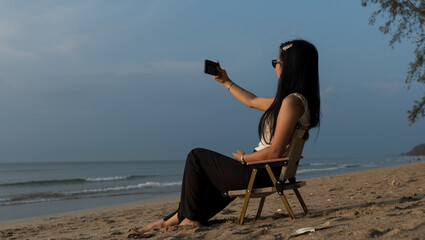 Woman taking a selfie on a beach chair with the sea and soft blue sky behind her.