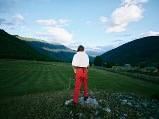 Obraz premium Hiker stands on a rocky ledge overlooking a green valley with towering mountains under a bright sky, red pants contrasting the serene countryside and peaks