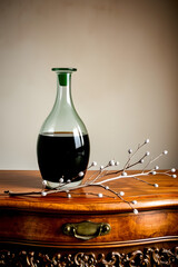 A refined glass decanter filled with dark green liquid, its translucent body reflecting subtle light, stands proudly on a rustic wooden dresser with a smooth, honey-brown finish