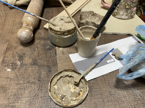 Messy high-angle shot of pottery studio workspace with jars of clay slip or glaze, brushes, a rolling pin, wooden shaping tools, and a small handmade clay bowl on a rustic wood surface
