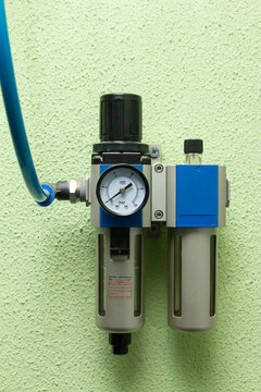 a compressed air filter and regulator unit (FRL) with A pressure gauge and two canisters, mounted on a light green textured industrial wall, with a blue tube connection on the left