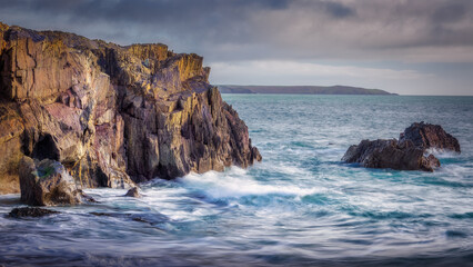 Strong waves crash around the base of a rugged cliff face on the coast. The sea rushes against the rocks.