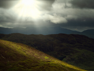 Sunlight shines through dark clouds onto a mountainous landscape in County Kerry, Ireland during the daytime.