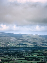 A distant view shows a valley with fields and towns below a mountain range. Clouds fill the sky above the landscape near the Coomloughra Horseshoe hiking trail.