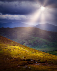 A scenic view of rolling hills and mountains under a dramatic sky. A bright ray of sunshine breaks through dark clouds, illuminating the landscape in County Kerry.