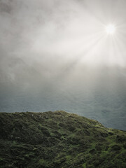 A rugged, green and rocky summit and a bright sun shining through the clouds, seen from the Horseshoe trail in Coomloughra, Ireland.