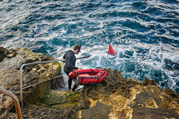 A spearfisherman in a wetsuit, holding a speargun and a red buoy with a flag, descends into the rough sea from a rocky shore.