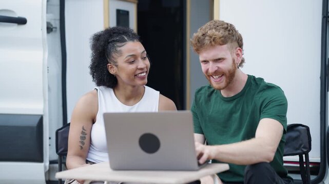 Happy couple enjoying their van life, sitting outside their camper van and using a laptop to plan their next travel destination
