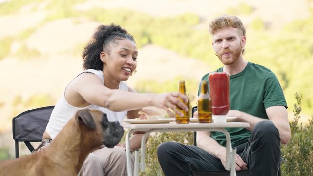 Young couple having a picnic with their boxer dog. Man and woman eating burgers and drinking beer outdoors, enjoying a romantic date
