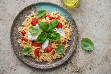 Plate of orzo pasta with mozzarella, roasted tomatoes and basil, horizontal shot on a beige granite...