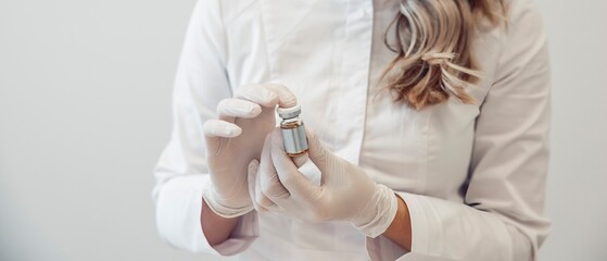 A female cosmetologist holds a vial of vaccine in a clinical setting. She wears gloves and a white coat, preparing for an injection.