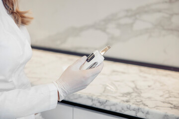 A female cosmetologist in a white coat holds a skincare device in a modern clinic. The countertop...