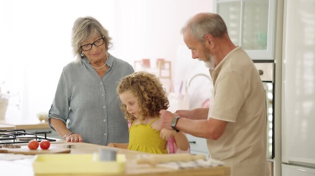 Loving grandfather helping his little granddaughter put on her pink backpack while grandmother watches smiling, getting her ready for school
