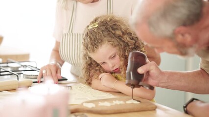 Cute curly-haired girl watching her grandfather pour chocolate frosting on homemade cookies. Family spending quality time together baking