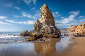 Majestic towering sea stack rock formations reflecting on wet sandy beach under clear blue sky with gentle ocean waves and distant rugged cliffs on sunny day