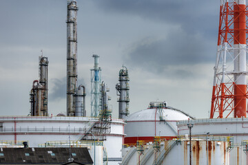 Fototapeta premium Sprawling industrial refinery with distillation columns and red-white lattice supported flue towering over facility's large storage tanks under bluish gray overcast sky.