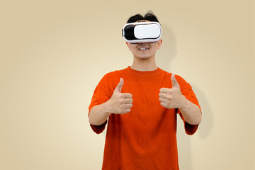 Happy young Asian man in an orange t-shirt wearing a VR headset and giving a double thumbs-up gesture, symbolizing approval, excellent experience studio background