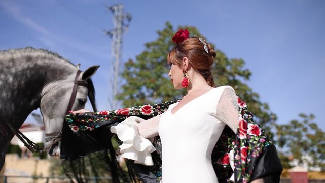 Elegant flamenco dancer in a traditional white dress and floral shawl caressing a stunning gray horse. Spanish culture and tradition concept