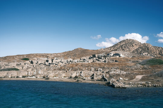 Clear coastal view of Delos island showing scattered ancient ruins on a rocky hillside, bright blue sky, and deep turquoise water below.