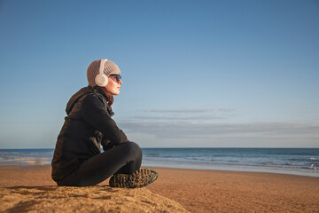 Woman sitting on a beach wearing headphones and winter jacket, enjoying the ocean view