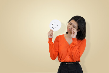 young Asian woman in an orange sweater rubbing her eyes as if crying, while holding a happy face emoji sign, symbolizing hidden sadness, emotional exhaustion, or masking deep sorrow 
