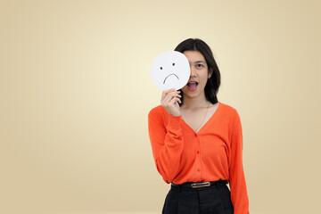 Smiling young Asian woman in an orange sweater, holding a sad face emoji sign to partially cover her face, symbolizing the emotional contradiction of masking inner sadness