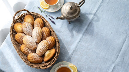 Basket of classic Madeleines dusted with sugar, served with lemon tea, overhead view.