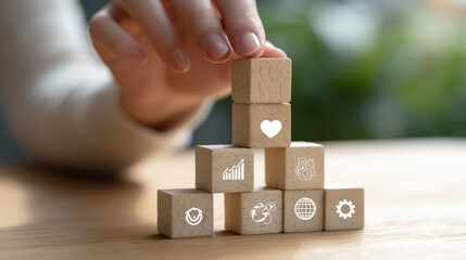 a person stacking wooden cubes into a pyramid shape, each cube has an icon: heart, gear, graph, globe, on a light wood table, natural daylight