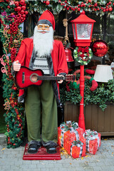 Festive Santa Claus statue holding a guitar, surrounded by Christmas decorations, gifts, greenery, and a bright red holiday lantern.