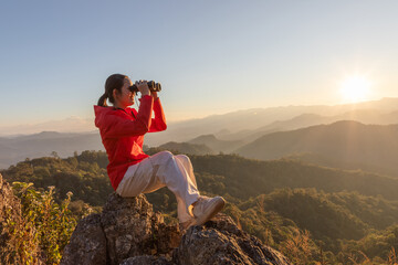 Cute girl hiker holding binoculars sitting on top of the rock mountain sunset sky background