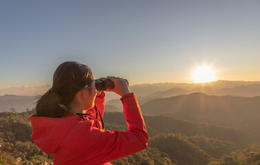 Cute girl hiker holding binoculars looking on top of the rock mountain sunset sky background