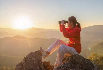 Cute girl hiker holding binoculars sitting on top of the rock mountain sunset sky background