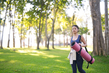 Asian adult woman carries yoga mat in lush park for outdoor fitness exercise wellness healthy lifestyle in serene green nature space for morning activity