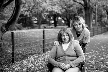 Elderly woman with grey hair in wheelchair on a walk in the park in fall weather with nurse or health care worker in blue uniform, both smiling happy