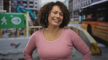 Hispanic woman in pink shirt with arms akimbo in front of building under renovation amidst urban barricades; confidence.
