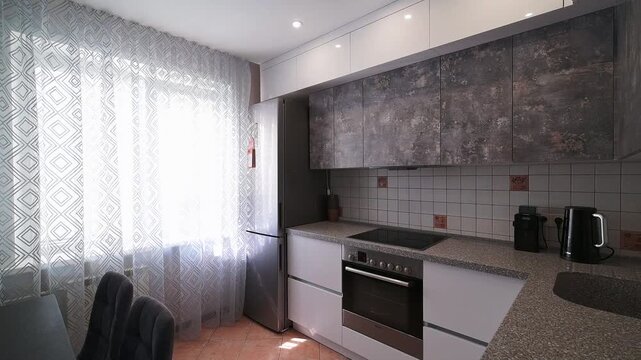 modern kitchen with dark dining table, geometric-patterned accent wall, white cabinetry, stainless steel appliances, pendant lights, sheer curtains, and tiled backsplash. Bright and stylish