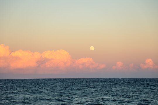 Crimson sunset skies glow above the open sea as the moon rises over gentle pink clouds, creating a serene, dreamlike coastal atmosphere ideal for tranquil nature backgrounds. - Powered by Adobe