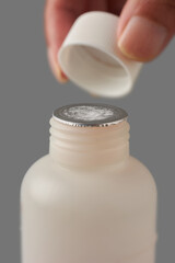 close-up of medicine bottle with intact foil safety seal, hand holding cap above container isolated neutral gray background, tamper evident packaging and pharmaceutical concept
