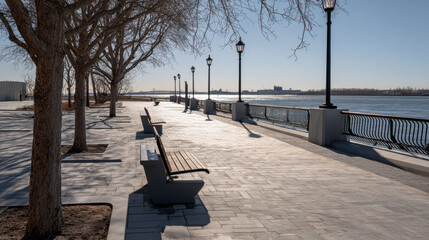 A serene view of a waterfront promenade featu benches and lampposts casting shadows along a paved path on a bright sunny day with a cityscape backdrop.