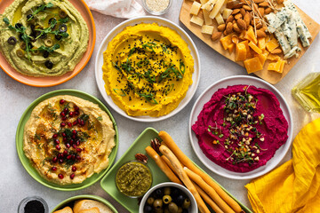 A variety of hummus flavors including turmeric, classic, and broccoli are served with fresh bread, nuts, and garnishes on a colorful charcuterie board, perfect for sharing