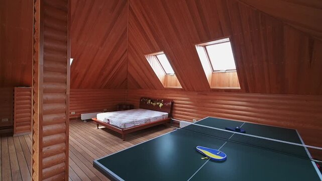 cozy wooden attic room features ping pong table, skylights, and bed. warm wooden walls and floor create  rustic atmosphere, while natural light enhances spacious feel