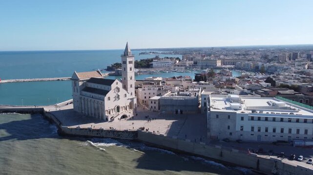 A panoramic coastal view of Trani shows the famous seaside cathedral overlooking turquoise waters, a long breakwater with lighthouses, and the historic old town bathed in bright Mediterranean sunlight