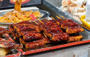 Grilled meat skewers on display at a food market in the evening with various other dishes nearby